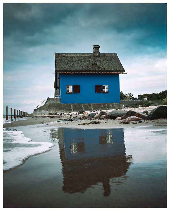 Ein Haus wie ein Gemälde – kräftiges Blau, umgeben von dramatischen Wolken, und seine Spiegelung im nassen Sand. Direkt an der Ostsee bei Heiligenhafen trotzt es Wind, Wellen und Wetter. Ein stiller Moment zwischen Ebbe und Flut, eingefangen in klarer Ruhe.