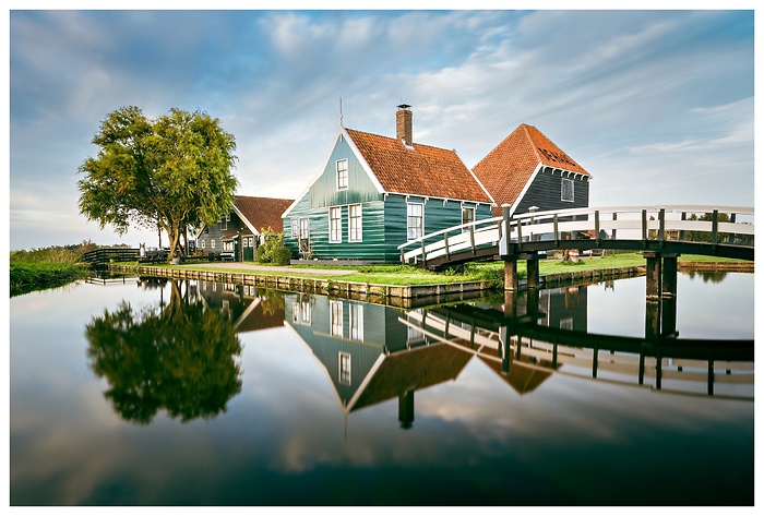 Traditionelle grüne Holzhäuser mit roten Ziegeldächern spiegeln sich in einem stillen Kanal. Eine weiße Holzbrücke und ein einzelner Baum vervollständigen die friedliche, niederländische Dorfromantik.