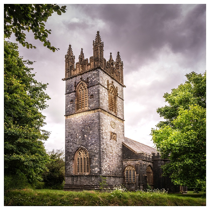 Dieses stimmungsvolle Bild fängt die majestätische St Mary's Church in Dartington, Devon, England, ein. Der markante, quadratische Kirchturm mit seinen gotischen Zinnen dominiert die Aufnahme und ragt vor einem dramatischen, bewölkten Himmel auf. Das graue Mauerwerk der Kirche, akzentuiert durch helle Fensterrahmen und Verzierungen, zeugt von ihrer langen Geschichte. Üppiges Grün umrahmt das Gebäude und verleiht der Szene eine friedliche und natürliche Atmosphäre. 
