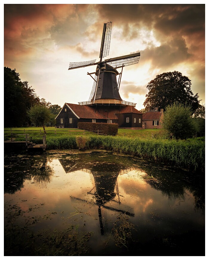 Eine historische Windm&uuml;hle erhebt sich ruhig in der flachen Landschaft, w&auml;hrend das warme Licht des sp&auml;ten Tages den Himmel f&auml;rbt. Im stillen Wasser davor spiegelt sich das Bauwerk und verleiht der Szene eine zeitlose, fast malerische Ruhe.