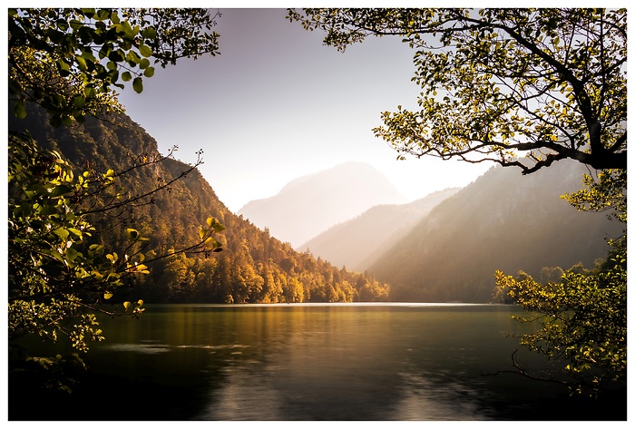 Im warmen Licht des fr&uuml;hen Abends liegt der See ruhig zwischen den Bergen. Herbstliche W&auml;lder spiegeln sich im Wasser, w&auml;hrend die Sonne die Landschaft in sanfte Goldt&ouml;ne taucht und einen Moment tiefer Stille entstehen l&auml;sst.