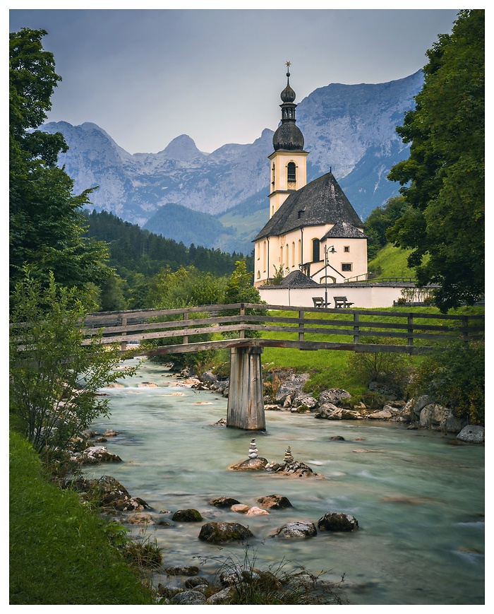 Die malerische Pfarrkirche St. Sebastian in Ramsau erhebt sich vor der Kulisse der Berchtesgadener Alpen. Ein klarer Gebirgsbach fließt sanft unter einer hölzernen Brücke hindurch und verstärkt den idyllischen Charme der Szenerie.