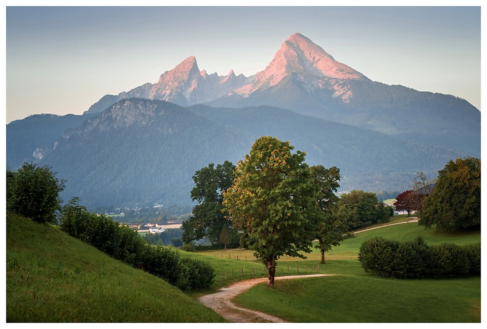 Die ersten Sonnenstrahlen des Tages tauchen den Watzmann in zartes Rosa. Vorne schlängelt sich ein idyllischer Weg durch sanfte Hügel und Wiesen – ein ruhiger Start in den Tag in den Berchtesgadener Alpen.