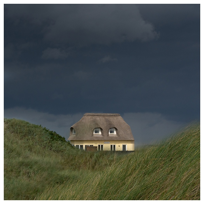 Ein Reetdachhaus inmitten der Dünen, umgeben von dramatischen, dunklen Gewitterwolken. Die Spannung liegt in der Luft – ein Moment vor dem Sturm, der gleichzeitig bedrohlich und wunderschön ist.
