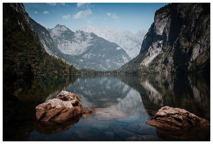 Ein ruhiger Bergsee liegt eingebettet zwischen steilen Felsw&auml;nden und bewaldeten H&auml;ngen. Die spiegelglatte Wasseroberfl&auml;che reflektiert die alpine Kulisse und verst&auml;rkt die zeitlose Ruhe dieses abgeschiedenen Ortes.