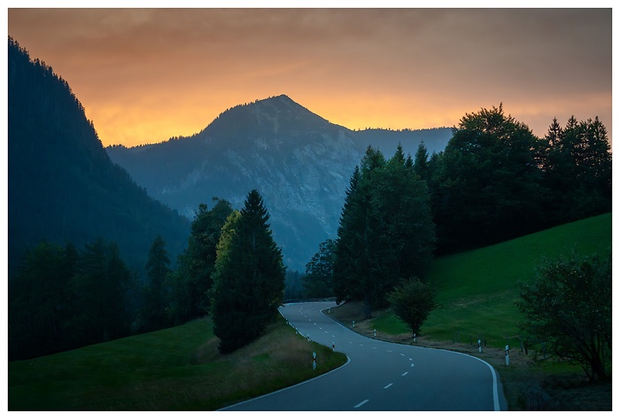 Eine geschwungene Straße führt durch die dunklen Wälder und sanften Hügel, während hinter den Bergen der Himmel in warmen Farben des Sonnenuntergangs erstrahlt.