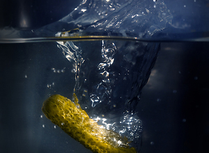 A green gherkin falls deep into a water glass.